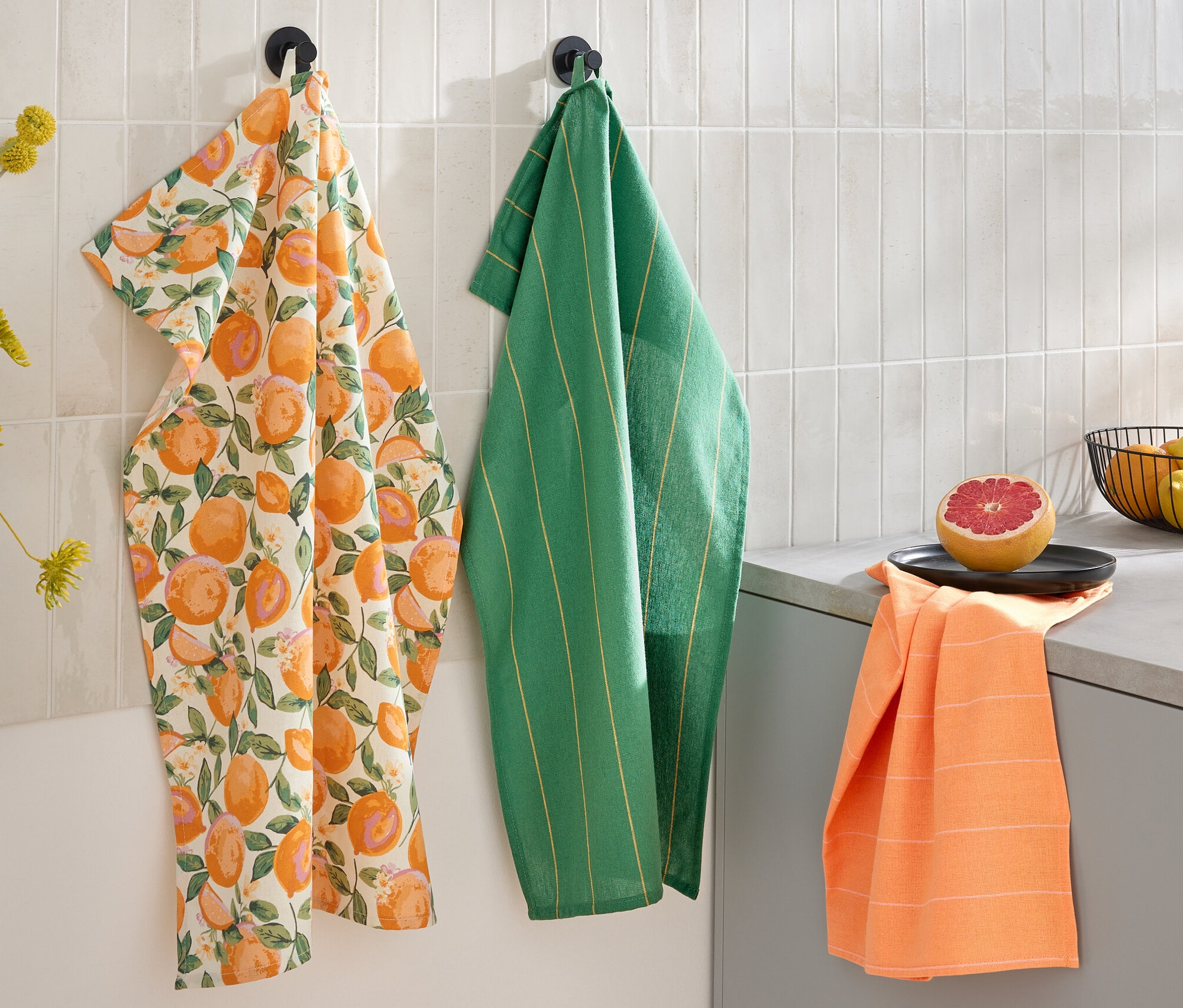 Drei Geschirrtücher hängen an der Wand: eines mit Zitrusdruck, eines grün gestreift und eines orangefarben. Auf der Arbeitsfläche liegen eine halbe Grapefruit und eine Schale mit Obst.