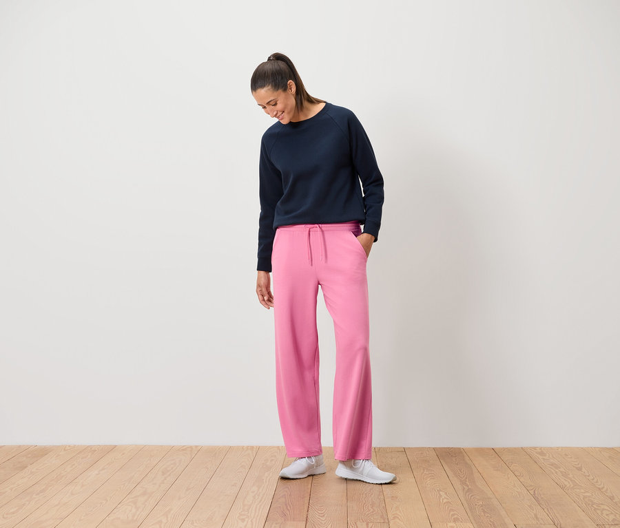 Eine Frau in einem marineblauen Sweatshirt und einer pinkfarbenen Hose steht auf einem Holzboden.