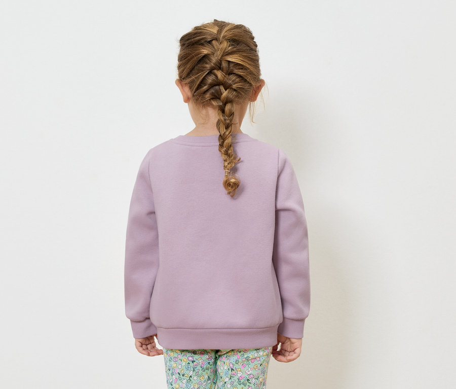 Mädchen von hinten mit Zopf, bekleidet mit einem lila Sweatshirt und einer geblümten Hose.