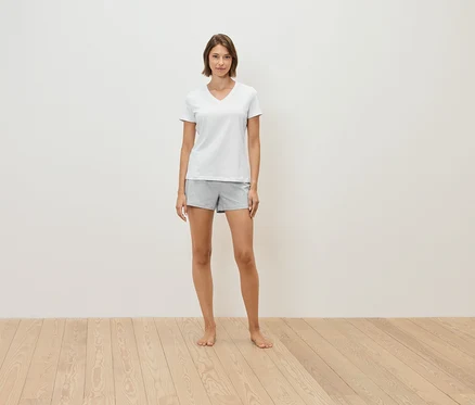 Frau steht barfuß auf einem Holzboden, trägt ein weißes T-Shirt und graue Shorts.