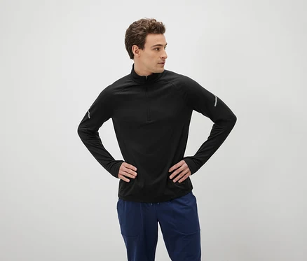 Mann mit braunen Haaren posiert mit den Händen in den Hüften, gekleidet in einem schwarzen Langarmshirt und blauen Hosen.