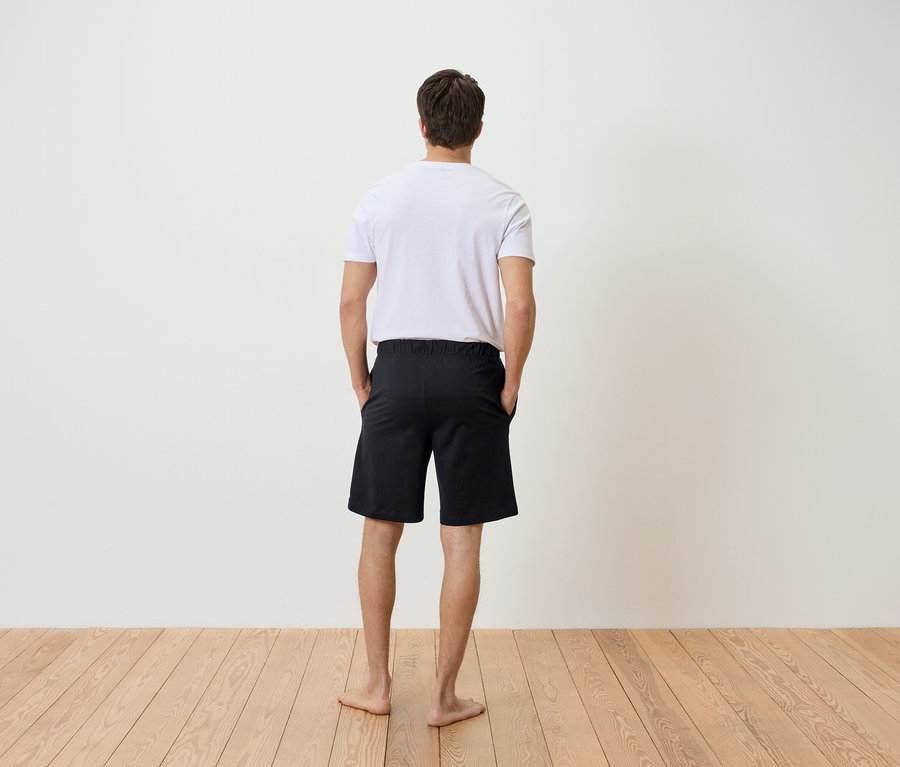 Mann von hinten, mit den Händen in den Hosentaschen einer schwarzen Shorts, dazu ein weißes T-Shirt, auf einem hellen Holzboden vor einer weißen Wand.