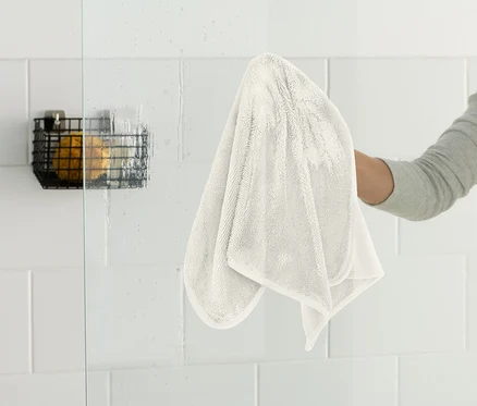Eine Person wischt eine Glasduschwand mit einem weißen Tuch ab. An der Wand hängt ein schwarzer Drahtkorb.