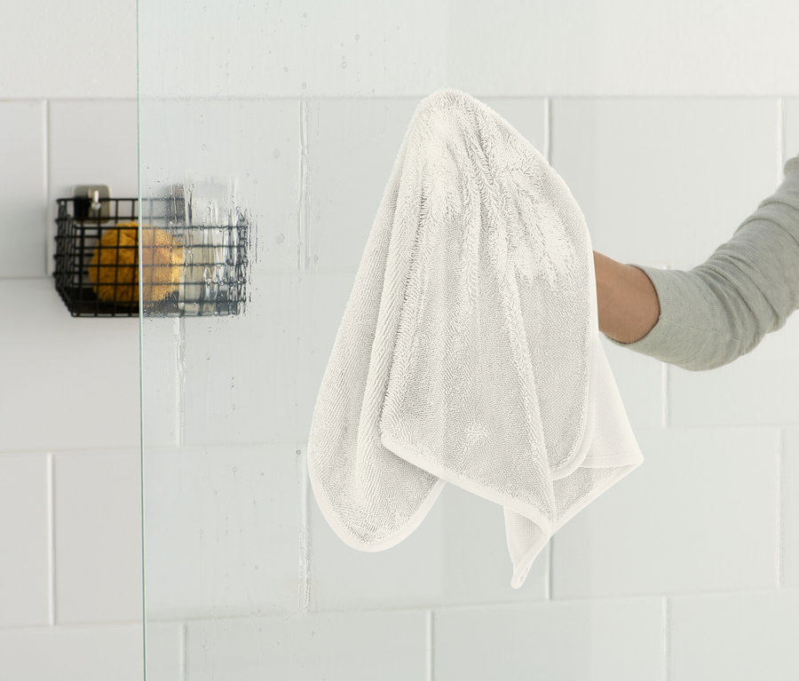 Eine Person wischt eine Glasduschwand mit einem weißen Tuch ab. An der Wand hängt ein schwarzer Drahtkorb.