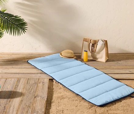 Eine hellblaue Strandmatte liegt auf einem Holzboden neben Sand, mit einem Strohhut, einer Sonnencremeflasche und einer Tragetasche in der Nähe.