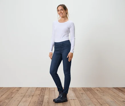 Eine Frau steht seitlich in Jeans, einem weißen Langarmshirt und Gummistiefeln vor einer weißen Wand und einem Holzboden.