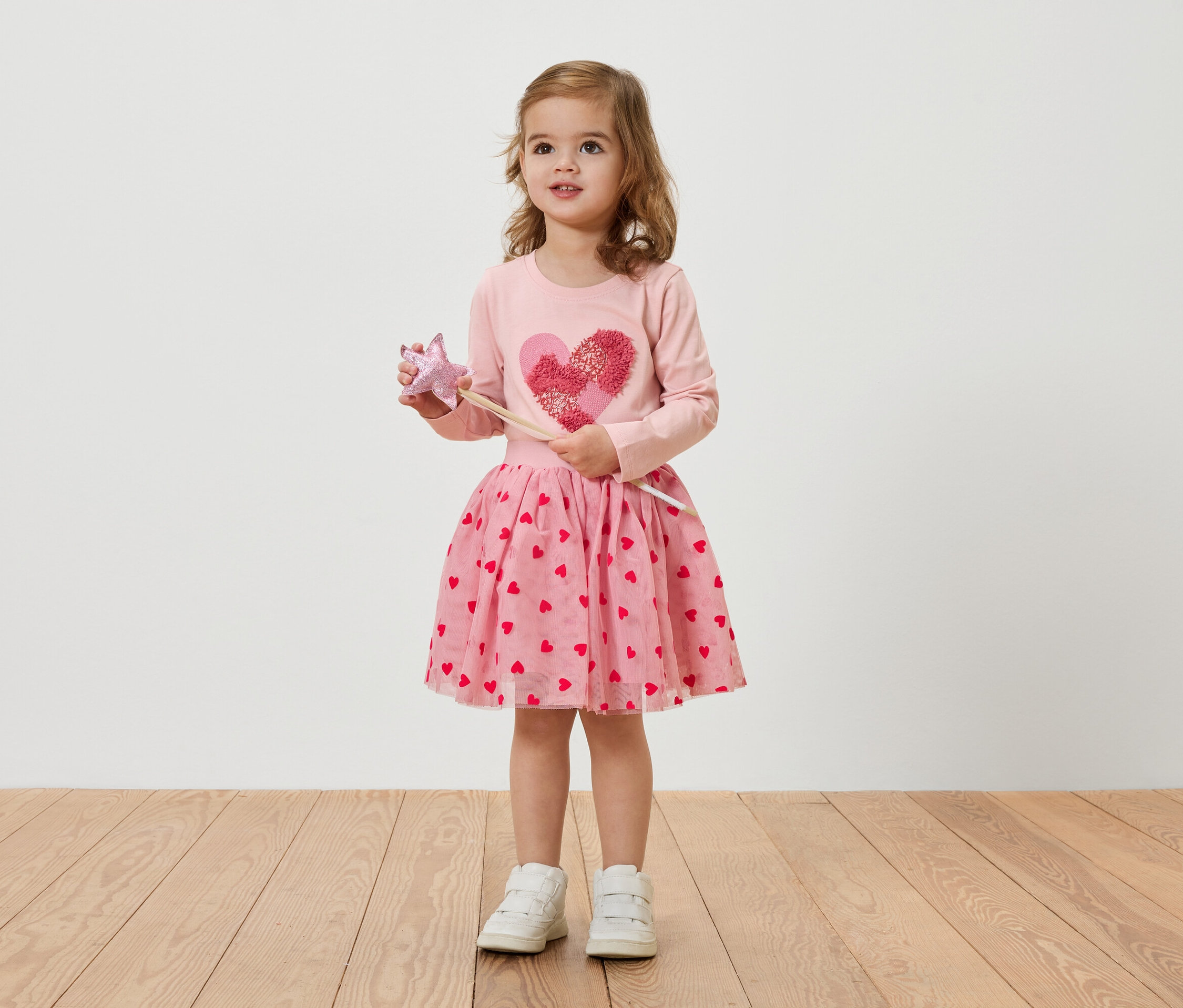 Ein kleines Mädchen mit blonden Haaren trägt ein rosa Herzchenkleid und hält einen Zauberstab.