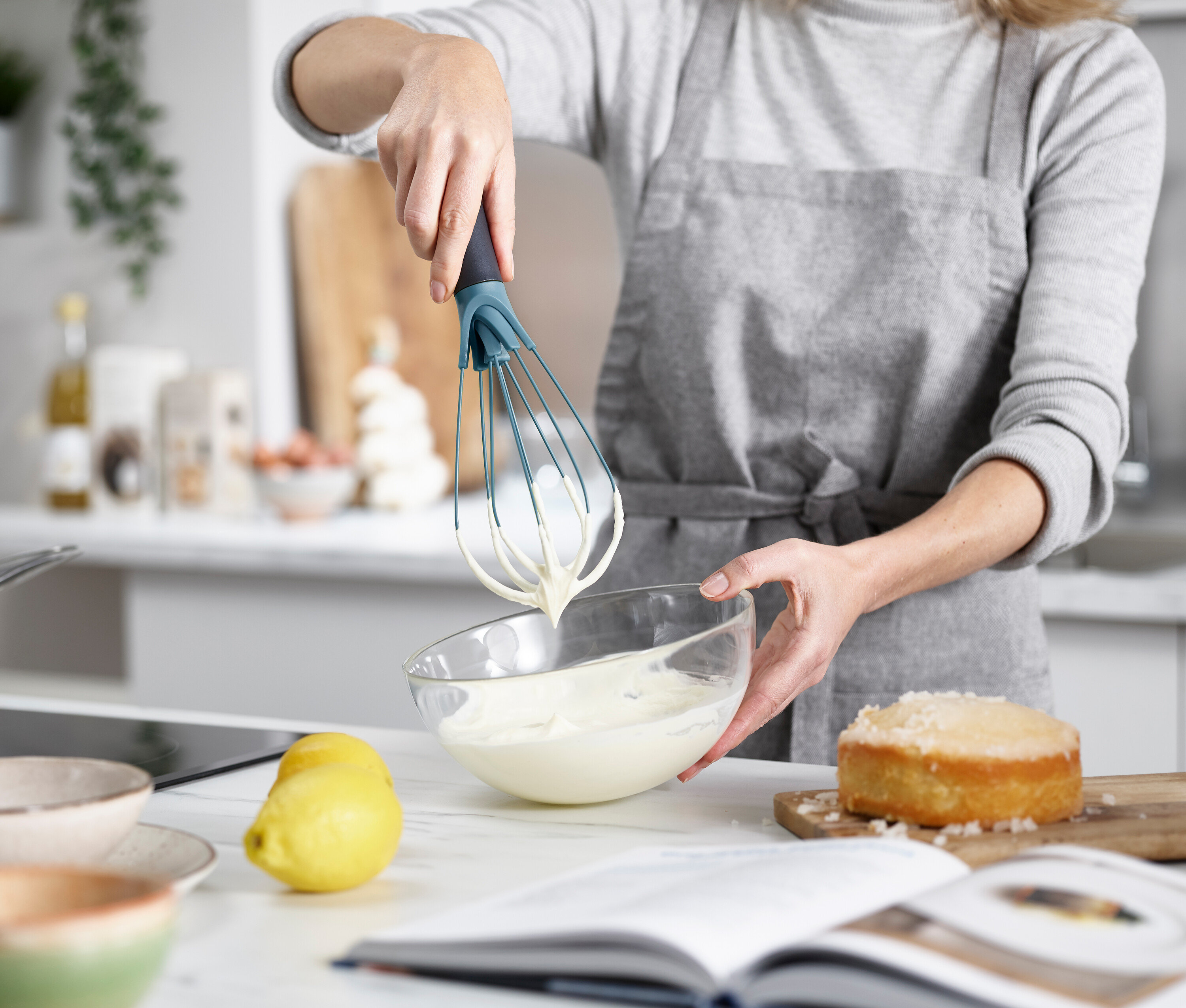 Eine Frau verrührt mit einem Schneebesen Teig in einer Glasschüssel, während ein Kuchen und eine Zitrone auf der Küchentheke stehen.