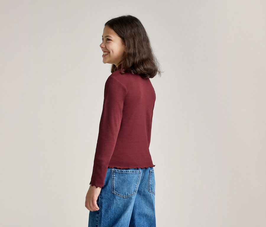 Ein lächelndes Mädchen mit langen, dunklen Haaren steht seitlich und trägt ein langärmeliges, weinrotes Oberteil mit Rüschensaum und blaue Jeans.