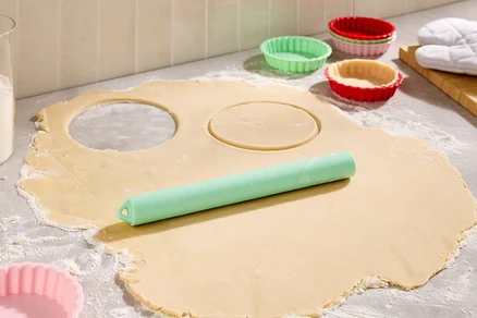 Auf einer bemehlten Arbeitsfläche liegt ausgerollter Teig mit zwei ausgestochenen Kreisen, ein grüner Teigroller und bunte Tarteförmchen.