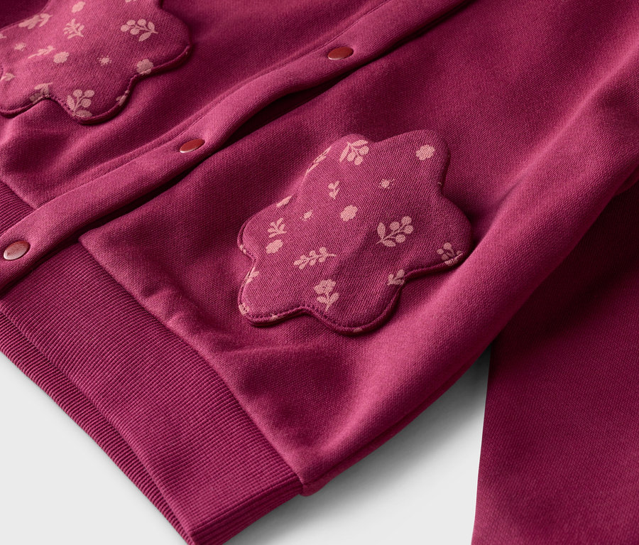 Nahaufnahme eines bordeauxroten Cardigans mit Blumenapplikationen und Knöpfen, sowie einer passenden Hose.