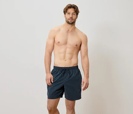 Mann steht barfuß vor einer weißen Wand und trägt eine marineblaue Badehose.