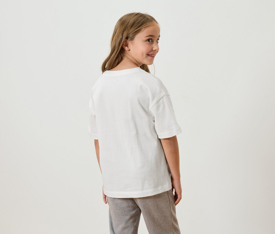 Ein junges Mädchen mit langen, braunen Haaren blickt über ihre Schulter. Sie trägt ein weißes T-Shirt und graue Jogginghosen.