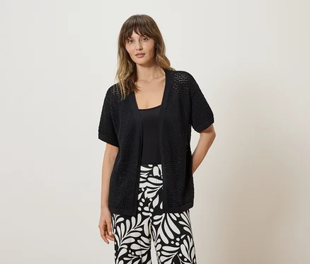 Frau trägt eine schwarze, kurzärmelige Strickjacke über einem schwarzen Top und schwarz-weiß gemusterte Hosen.
