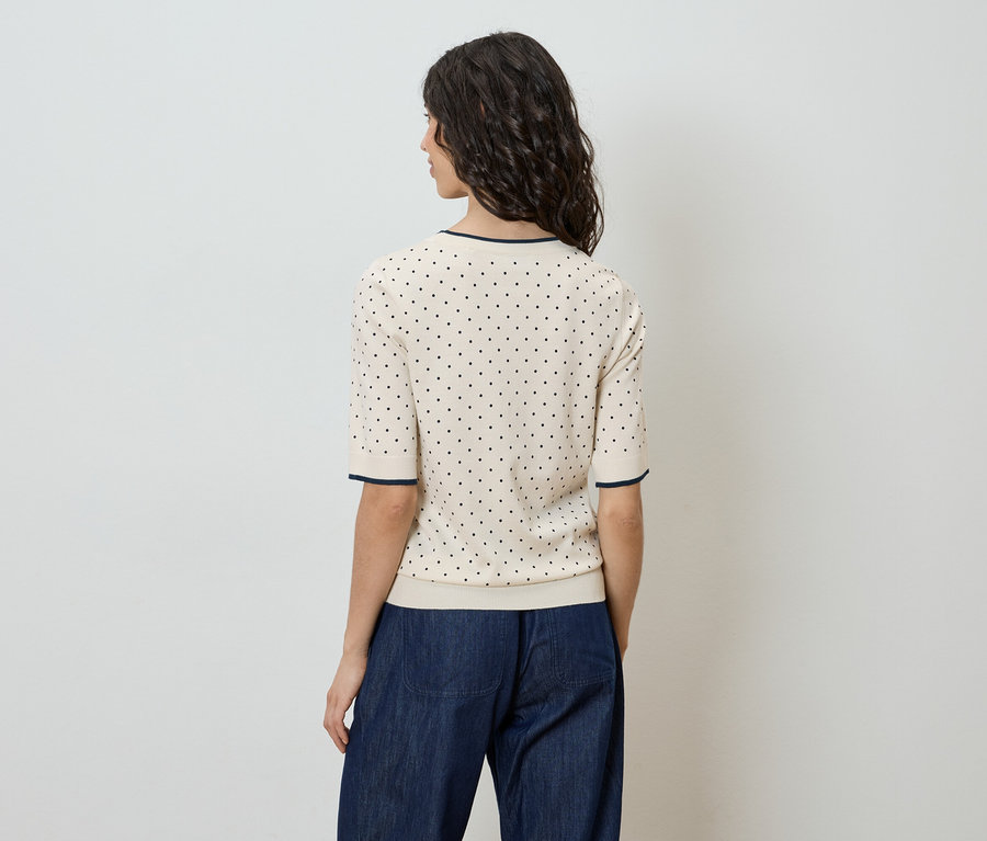 Rückansicht einer Frau mit gewelltem, dunklem Haar, die ein kurzärmeliges, helles Oberteil mit schwarzen Punkten und eine dunkle Jeans trägt.