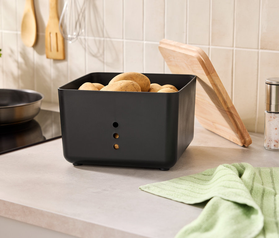 Auf einer Küchentheke steht ein schwarzer Kartoffelbehälter mit Kartoffeln, daneben ein Schneidebrett, Gewürzmühlen und ein Geschirrtuch.
