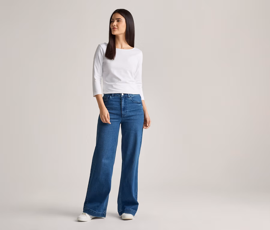 Frau mit langem, dunklem Haar, trägt ein weißes Langarmshirt, weite, blaue Jeans und weiße Schuhe vor einem hellen Hintergrund.