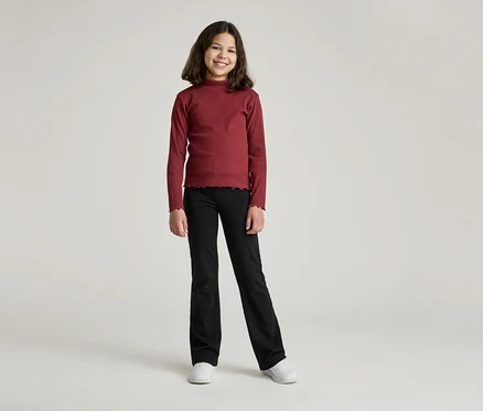 Ein junges Mädchen mit braunen, schulterlangen Haaren lächelt in einem bordeauxroten Langarmshirt und einer schwarzen Hose mit Schlag.
