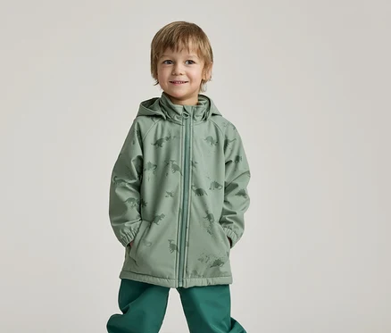 Ein lächelnder Junge mit Händen in den Taschen trägt eine grüne Regenjacke mit Dinosaurier-Muster und grüne Hosen vor einem hellen Hintergrund.