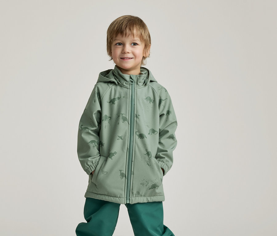 Ein lächelnder Junge mit Händen in den Taschen trägt eine grüne Regenjacke mit Dinosaurier-Muster und grüne Hosen vor einem hellen Hintergrund.
