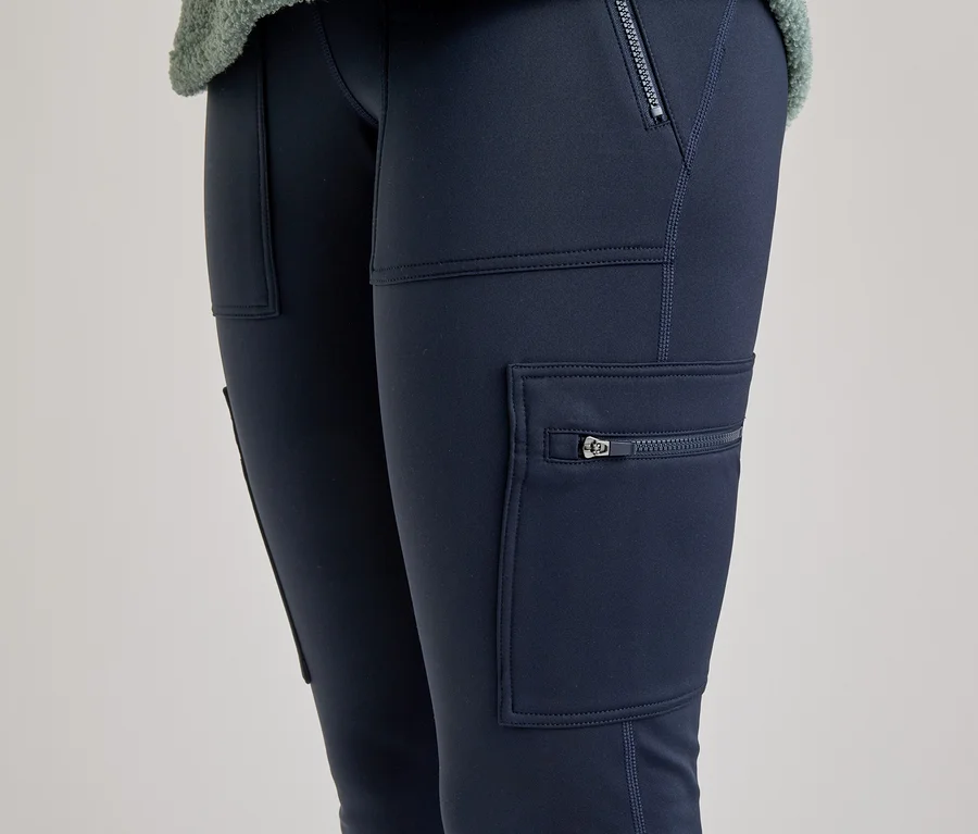 Nahaufnahme der Beine einer Person, die dunkelblaue Cargo-Leggings mit Reißverschlusstaschen und aufgesetzten Taschen trägt. Ein Teil eines hellgrünen, flauschigen Oberteils ist oben sichtbar.