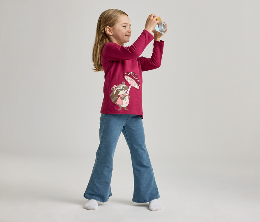 Mädchen mit pinkem Shirt mit Waschbärmotiv und blauen Jeans posiert mit Spielzeugkamera vor hellem Hintergrund.
