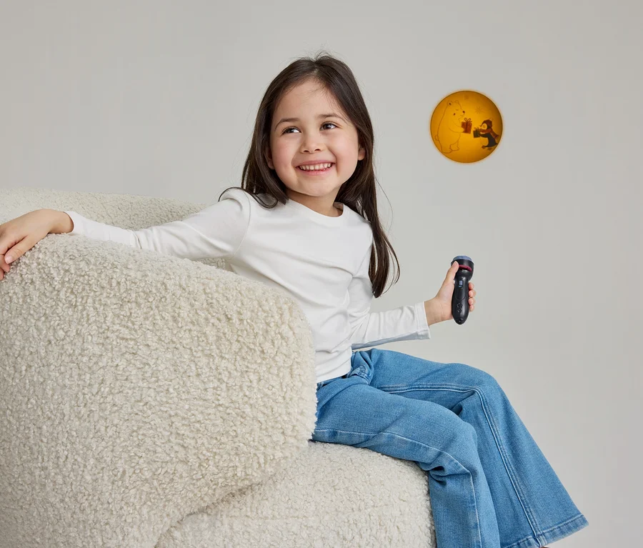 Ein lächelndes junges Mädchen mit langen dunklen Haaren sitzt in einem cremefarbenen, flauschigen Sessel. Sie trägt ein weißes Langarmshirt und blaue Jeans, hält einen schwarzen Spielzeugprojektor in der rechten Hand und blickt auf eine kreisförmige, gelb-orangefarbene Projektion an der Wand, die einen Eisbären und eine Person beim Geschenkeaustausch zeigt.