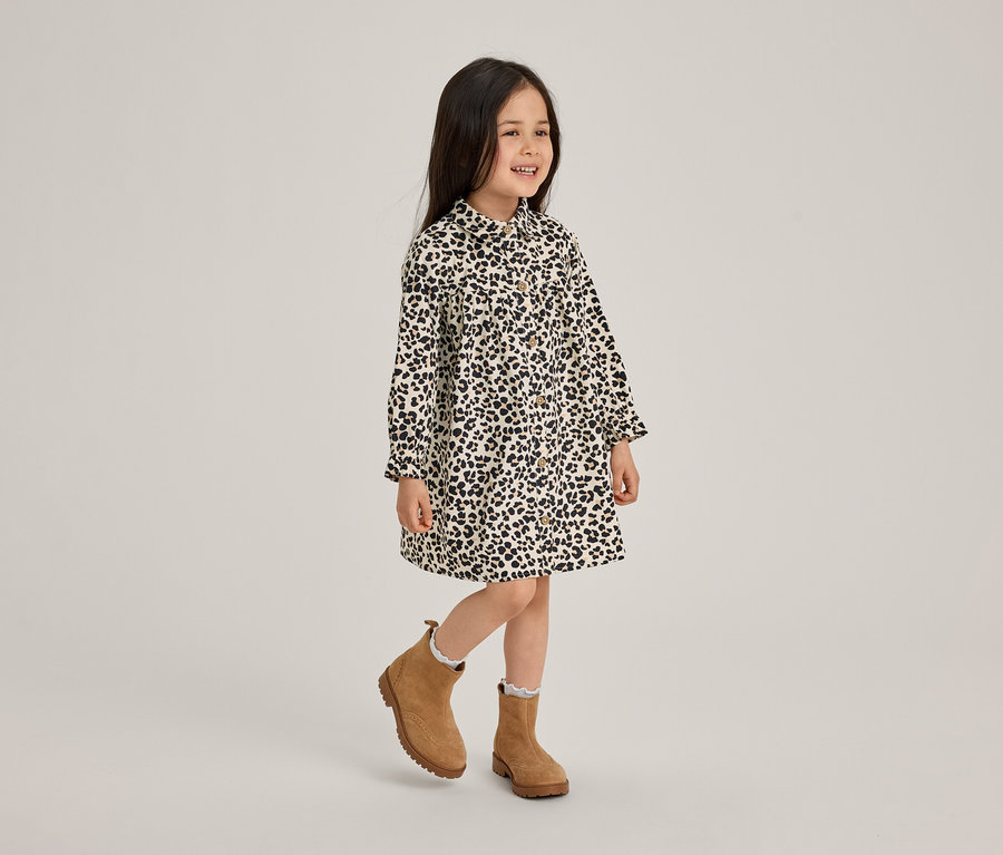 Ein lächelndes kleines Mädchen in einem schwarz-weiß gemusterten Kleid und braunen Stiefeletten steht auf einem Bein.