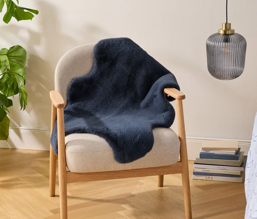 Beiger Sessel mit einem dunkelblauen Kunstfell-Überwurf, daneben eine große Monstera-Pflanze in einem geflochtenen Korb. Ein Stapel Bücher liegt am Boden, darüber hängt eine gerippte Glaslampe. Hellblaue gemusterte Bettwäsche ist rechts sichtbar.