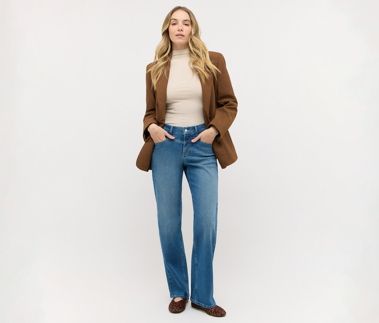 Frau mit langen blonden Haaren steht, trägt einen braunen Blazer über einem beigen Oberteil, blaue Jeans und Leoparden-Ballerinas, Hände in den Taschen.