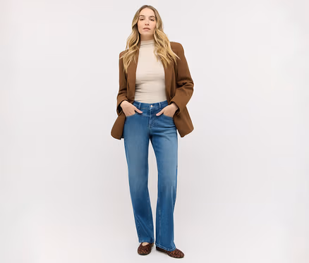 Frau mit langen blonden Haaren steht, trägt einen braunen Blazer über einem beigen Oberteil, blaue Jeans und Leoparden-Ballerinas, Hände in den Taschen.