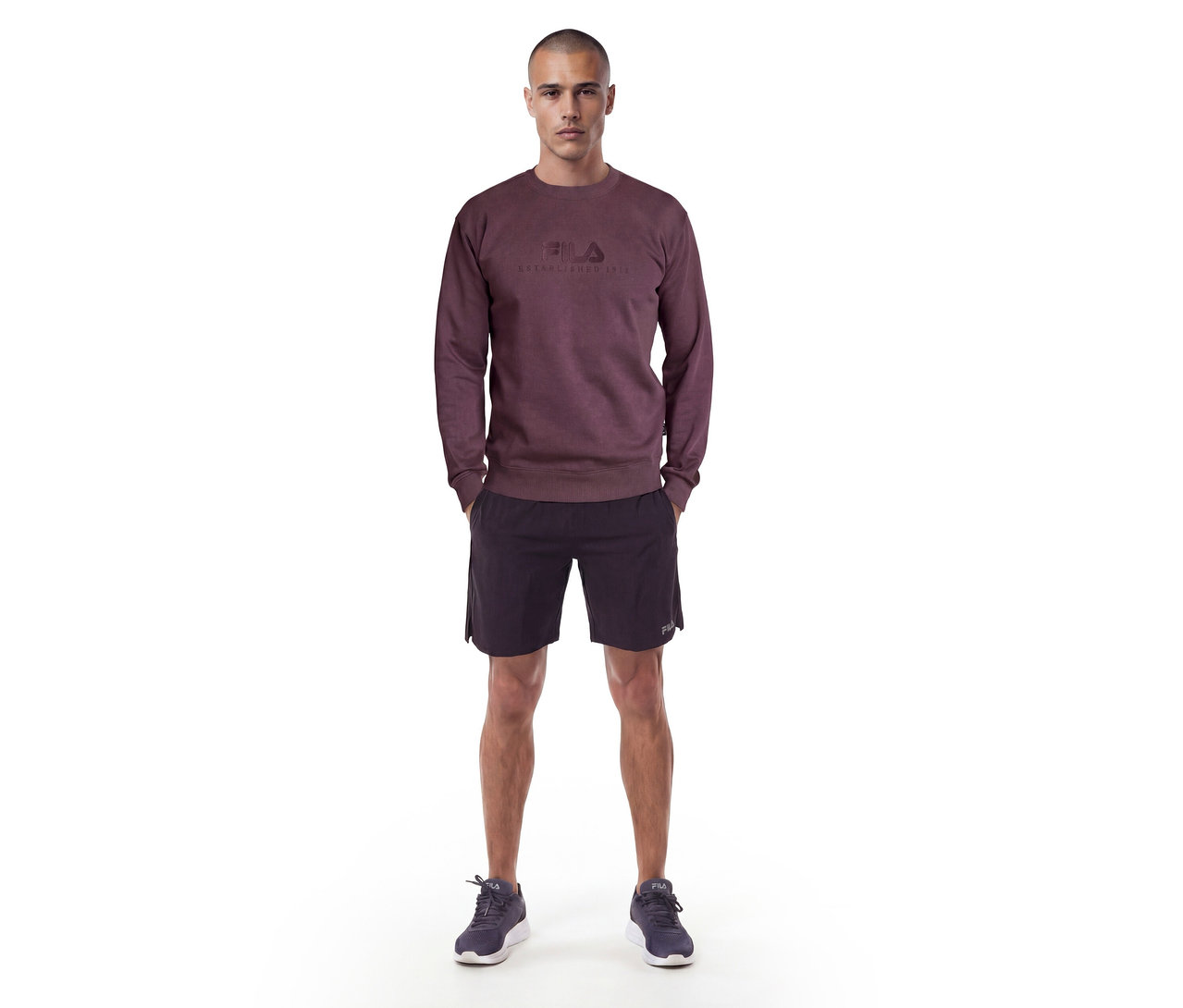 Mann steht in einem dunkelroten Sweatshirt mit Fila-Logo, schwarzen Shorts und dunklen Turnschuhen.