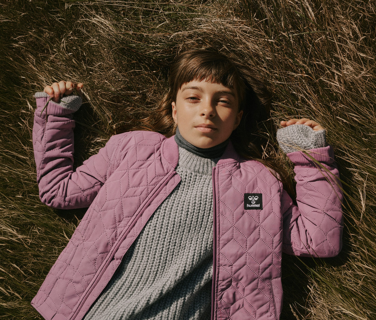 Ein junger Mensch liegt im trockenen Gras und trägt eine rosa gesteppte hummel® HMLSOBI Thermo-Set Jacke über einem grauen Strickpullover, blickt in die Kamera.