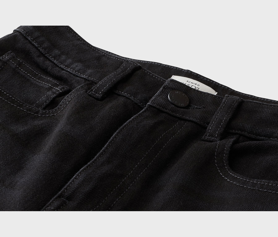Nahaufnahme einer schwarzen Jeans mit vorderen Taschen, Knopf, Gürtelschlaufen und einem teilweise sichtbaren weißen Tchibo-Etikett.