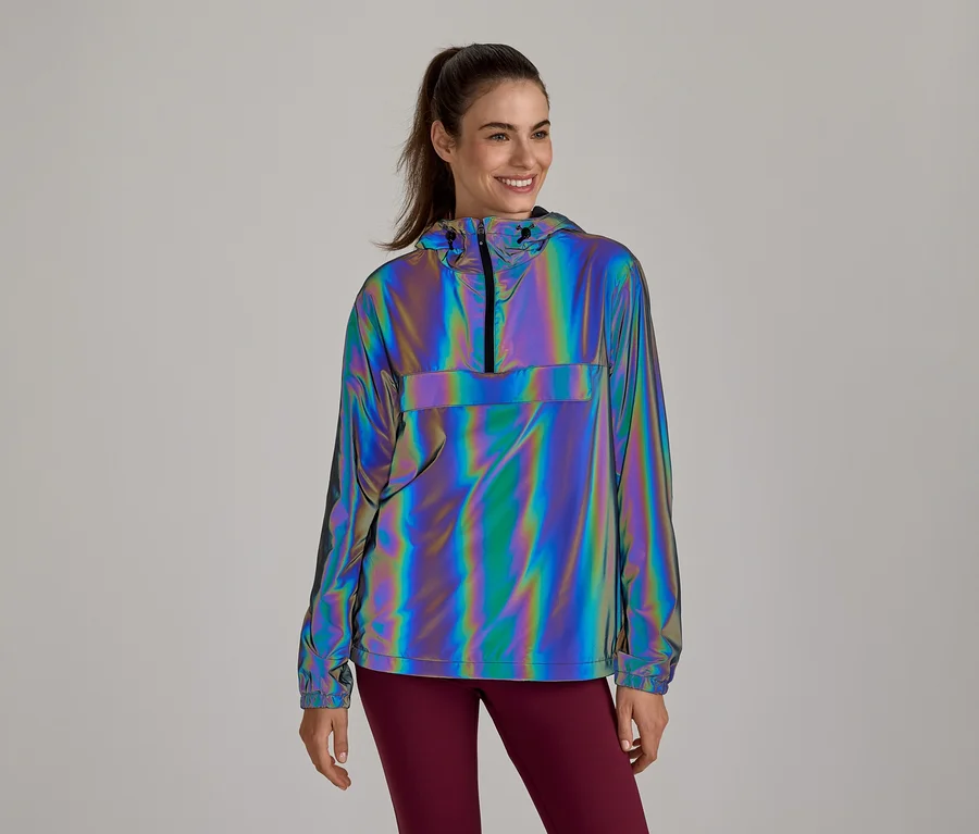 Lächelnde Frau trägt eine reflektierende, irisierende Anorak-Jacke mit Kapuze und halbem Reißverschluss, kombiniert mit dunkelroten Leggings.