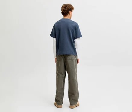 Eine Person steht mit dem Rücken zum Betrachter und trägt ein Jack&Jones double layered Shirt, bestehend aus einem blauen Kurzarmshirt über einem weißen Langarmshirt, dazu olivgrüne Hosen und hellbraune Schuhe.