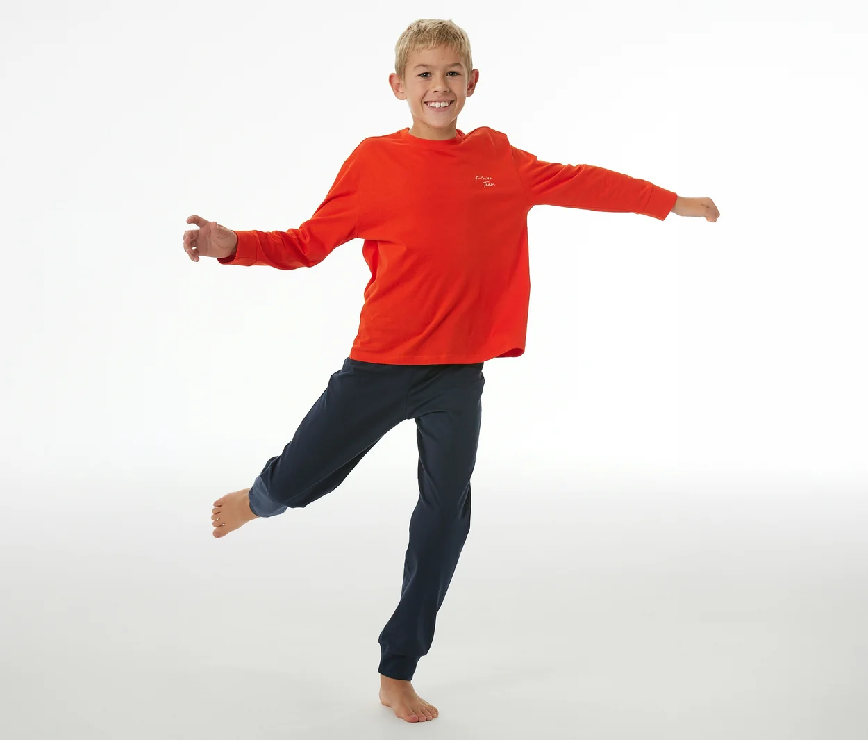 Lächelnder Junge in rotem SCHIESSER Kinder-Pyjama-Oberteil und dunkelblauer Pyjama-Hose mit Bündchen, der auf einem Bein balanciert und die Arme ausstreckt.