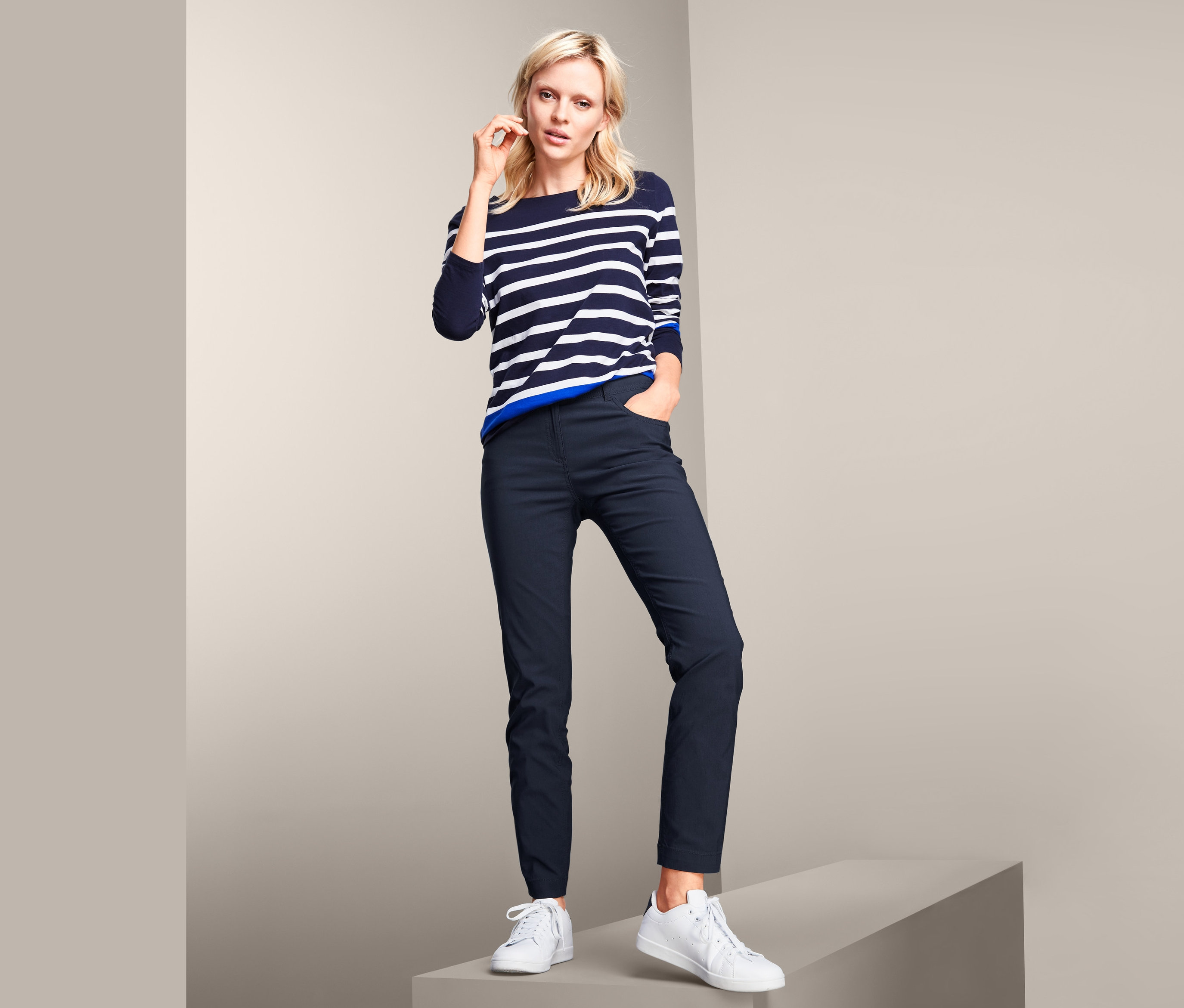 Blonde Frau posiert auf einer grauen Plattform in einem gestreiften Pullover und mit einer Hand in der Tasche ihrer Bengalin-Hose.