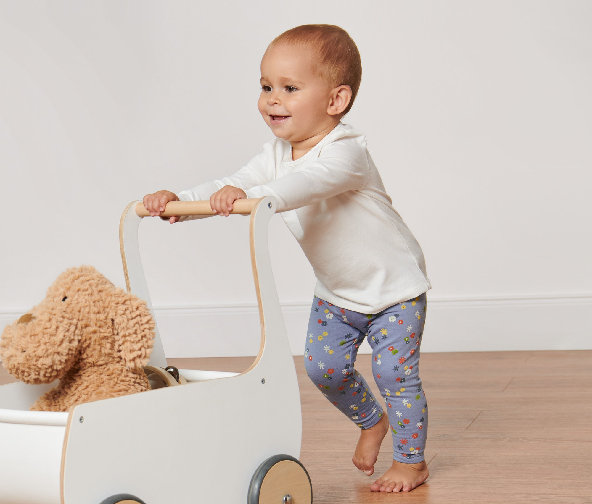 Ein Baby schiebt einen weißen Wagen mit einem Plüschhund und trägt lila Baby-und-Kleinkind-Leggings.