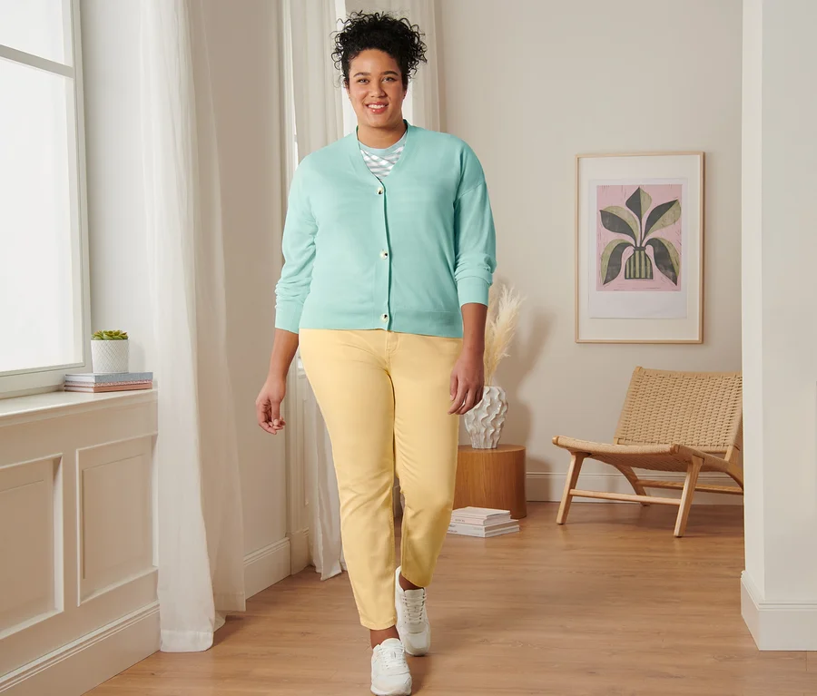 Eine Frau geht und trägt ein mintfarbenes, gestreiftes T-Shirt, einen Cardigan mit V-Ausschnitt, eine gelbe Slimfit-Jeans und Veloursleder-Sneaker.
