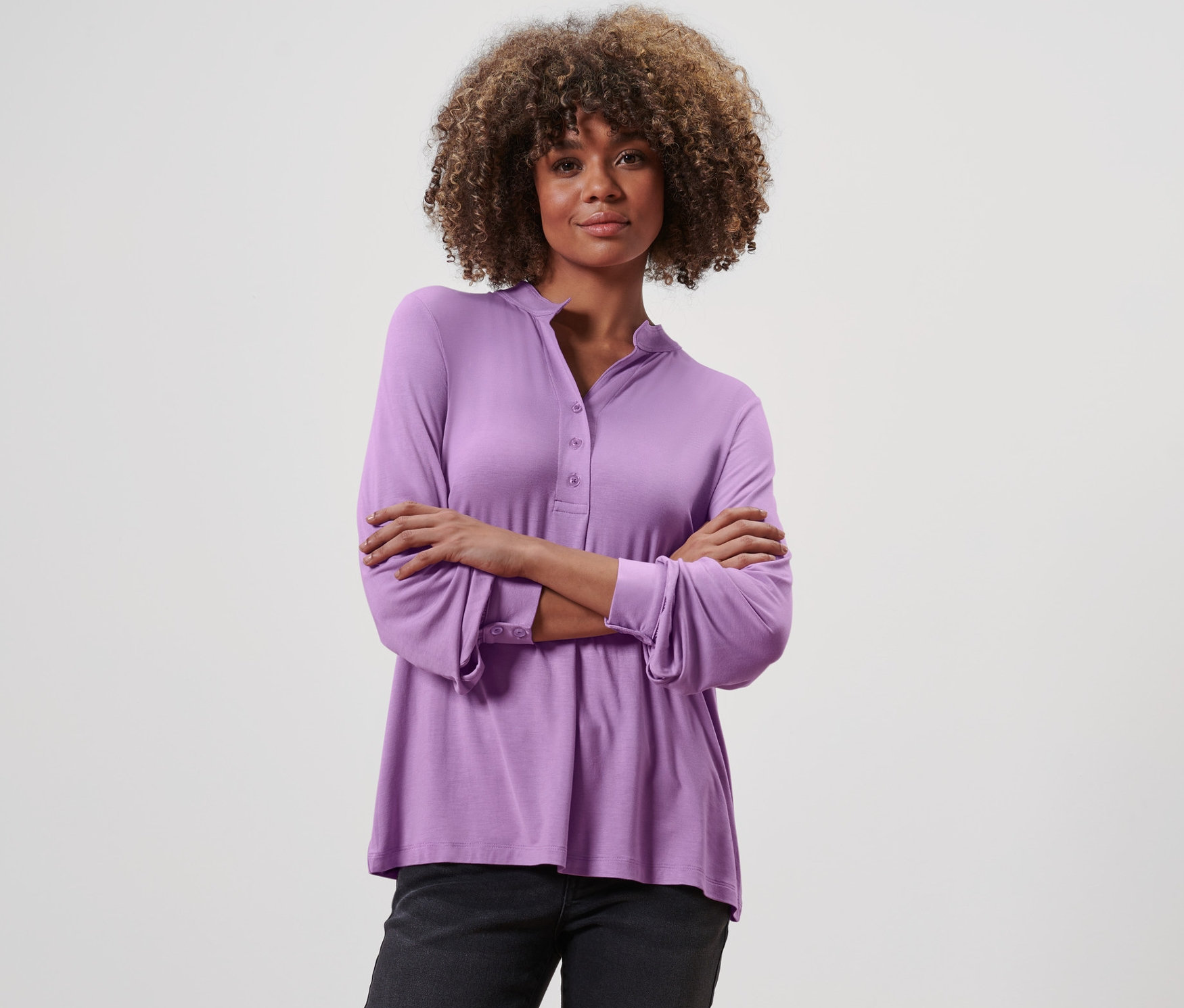 Frau posiert mit verschränkten Armen in einem fliederfarbenen Blusenshirt mit Knopfleiste und dunkler Hose.