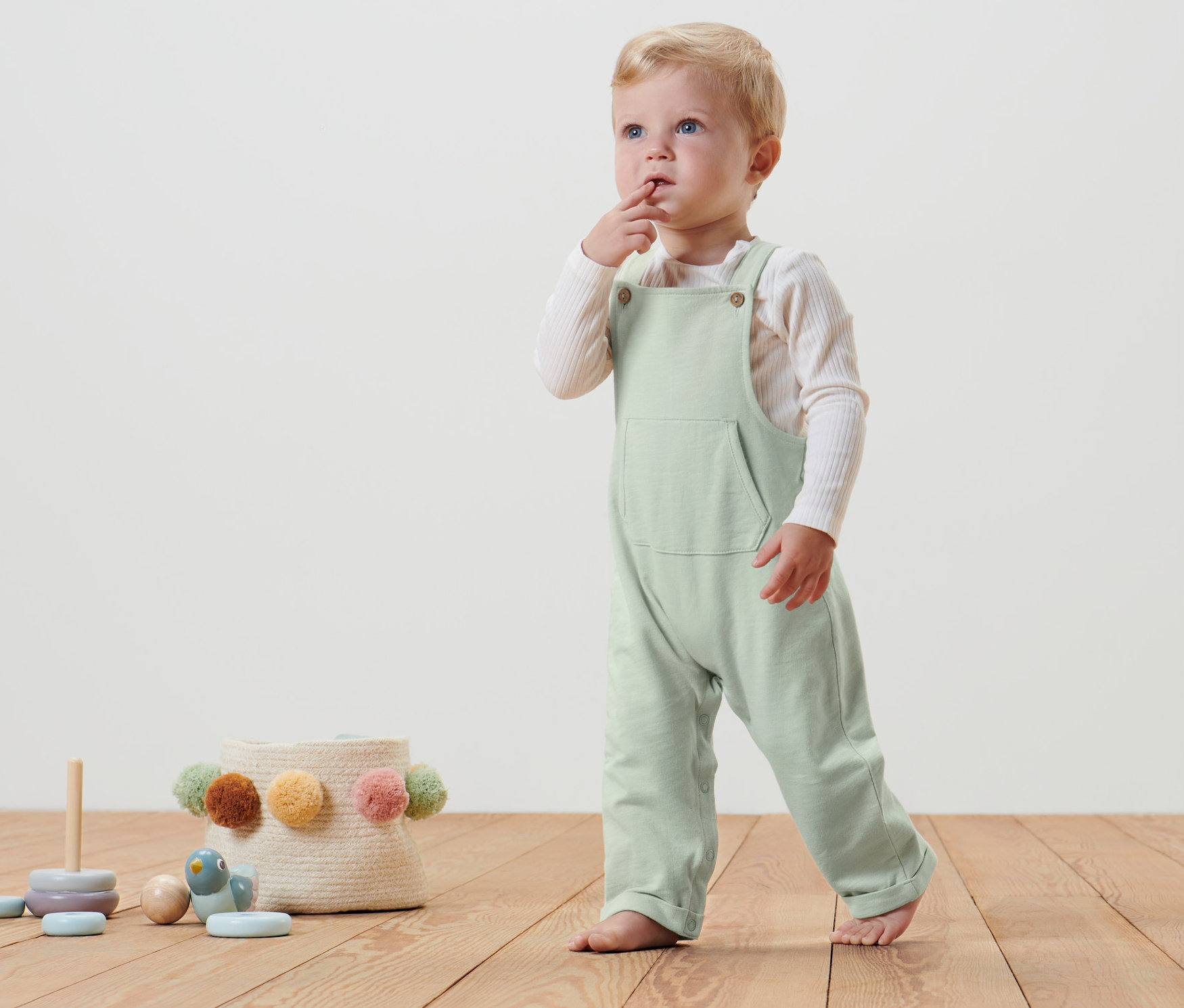 Ein Baby in einer Baby-Latzhose steht auf einem Holzboden und lutscht an seinem Finger.