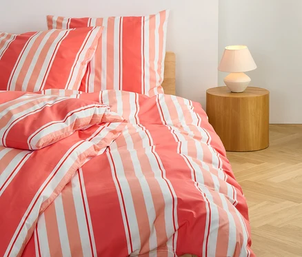 Detailansicht von koralle gestreifter Renforcé-Bettwäsche in Normalgröße und Übergröße auf einem Bett.
