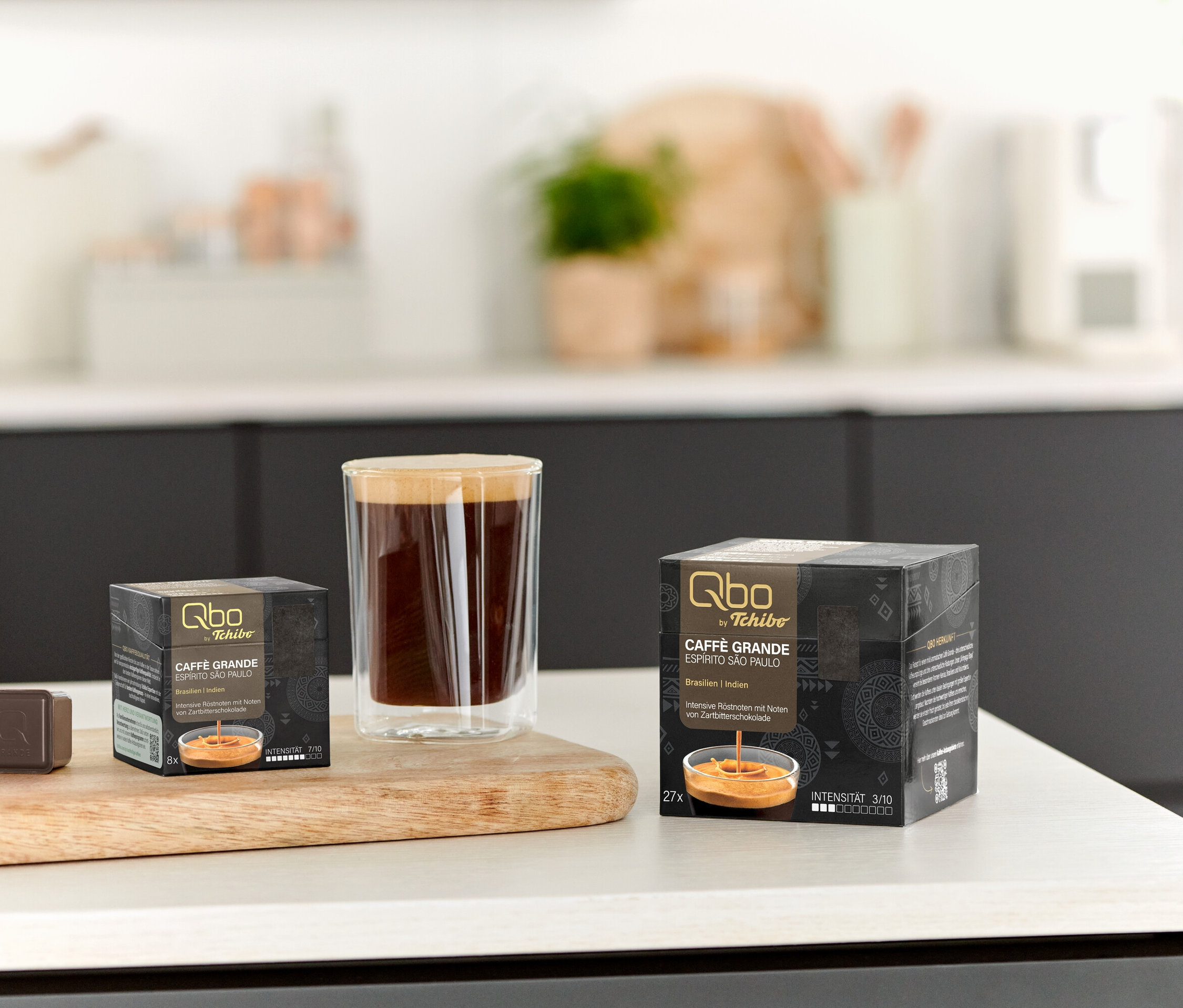 Auf einem Tisch stehen eine Tasse Kaffee und Packungen Qbo Caffè Grande ESPÍRITO SÃO PAULO mit 8 und 27 Kapseln.