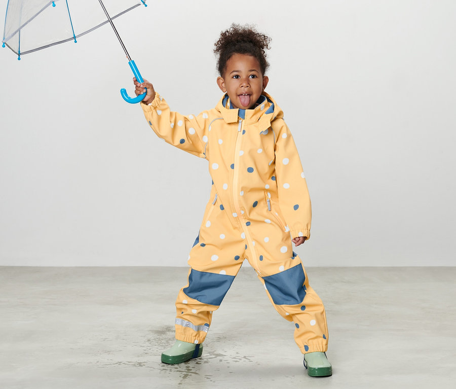 Ein Kind hält einen durchsichtigen Regenschirm und trägt einen Kleinkind-Softshell-Einteiler mit Punkte-Alloverprint.
