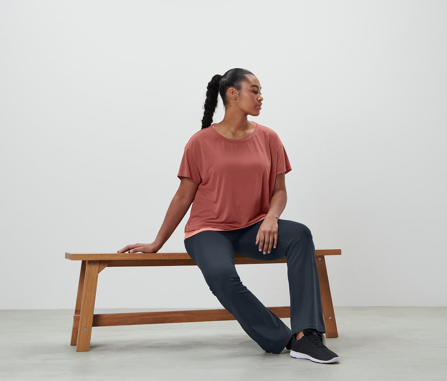 Frau sitzt auf einer Bank und trägt eine Bodyforming-Sporthose und ein 2-in-1-Kurzarm-Sportshirt in Terrakotta.