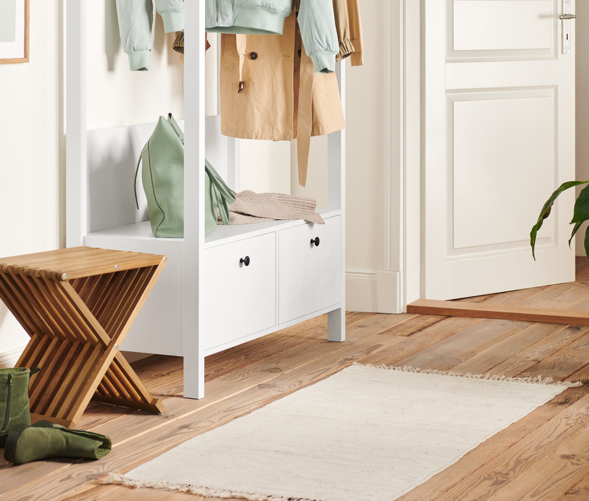 Im Flur stehen eine weiße Garderobe mit großen Schubladen und ein Hocker mit Klappfunktion. Auf der Garderobe liegen eine grüne Handtasche und ein Strickpullover.