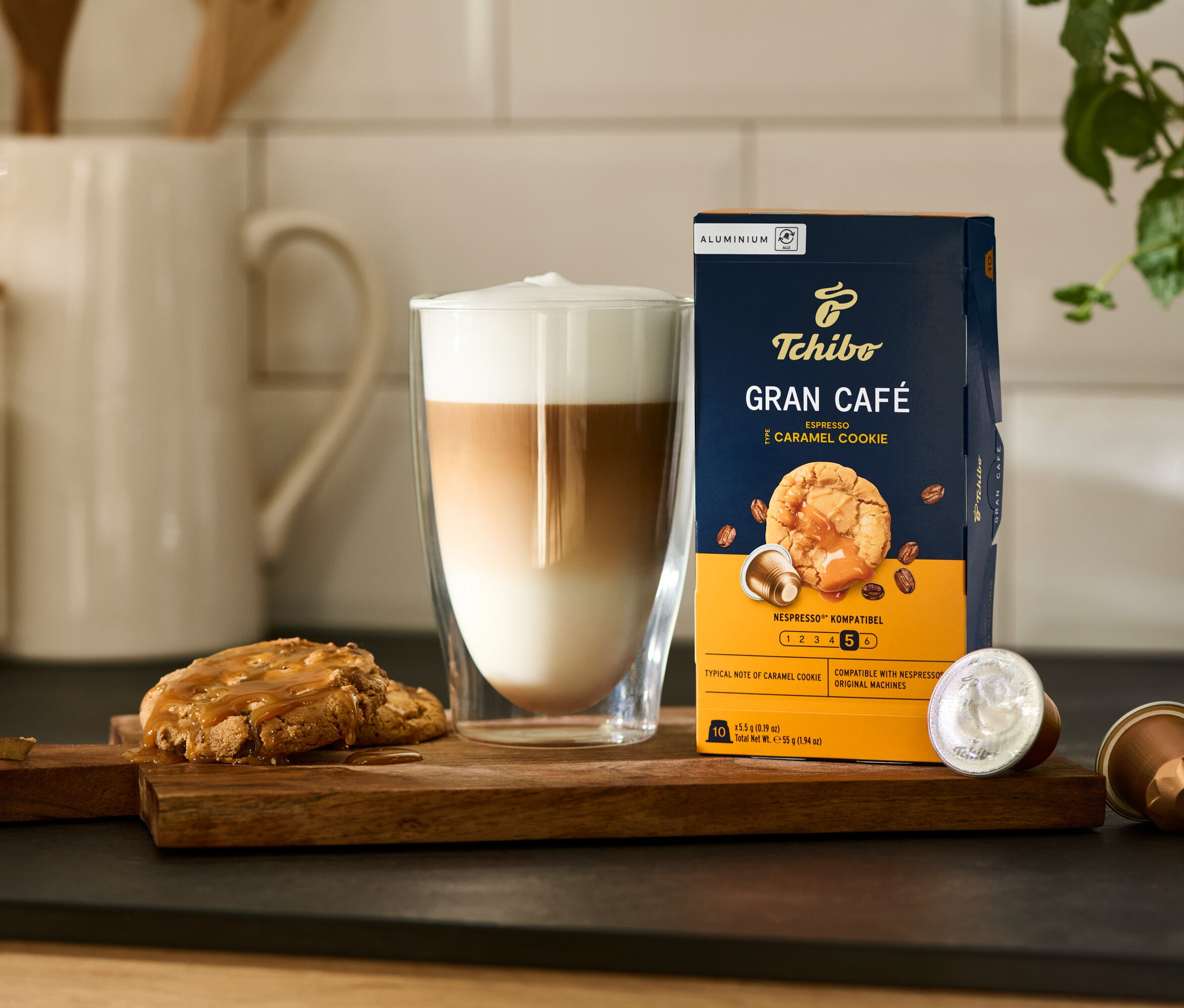 Auf einem Holzbrett stehen ein Glas Latte Macchiato, ein Keks und eine Packung Tchibo Gran Café Flavoured Espresso Caramel Cookie - 10 Kapseln.