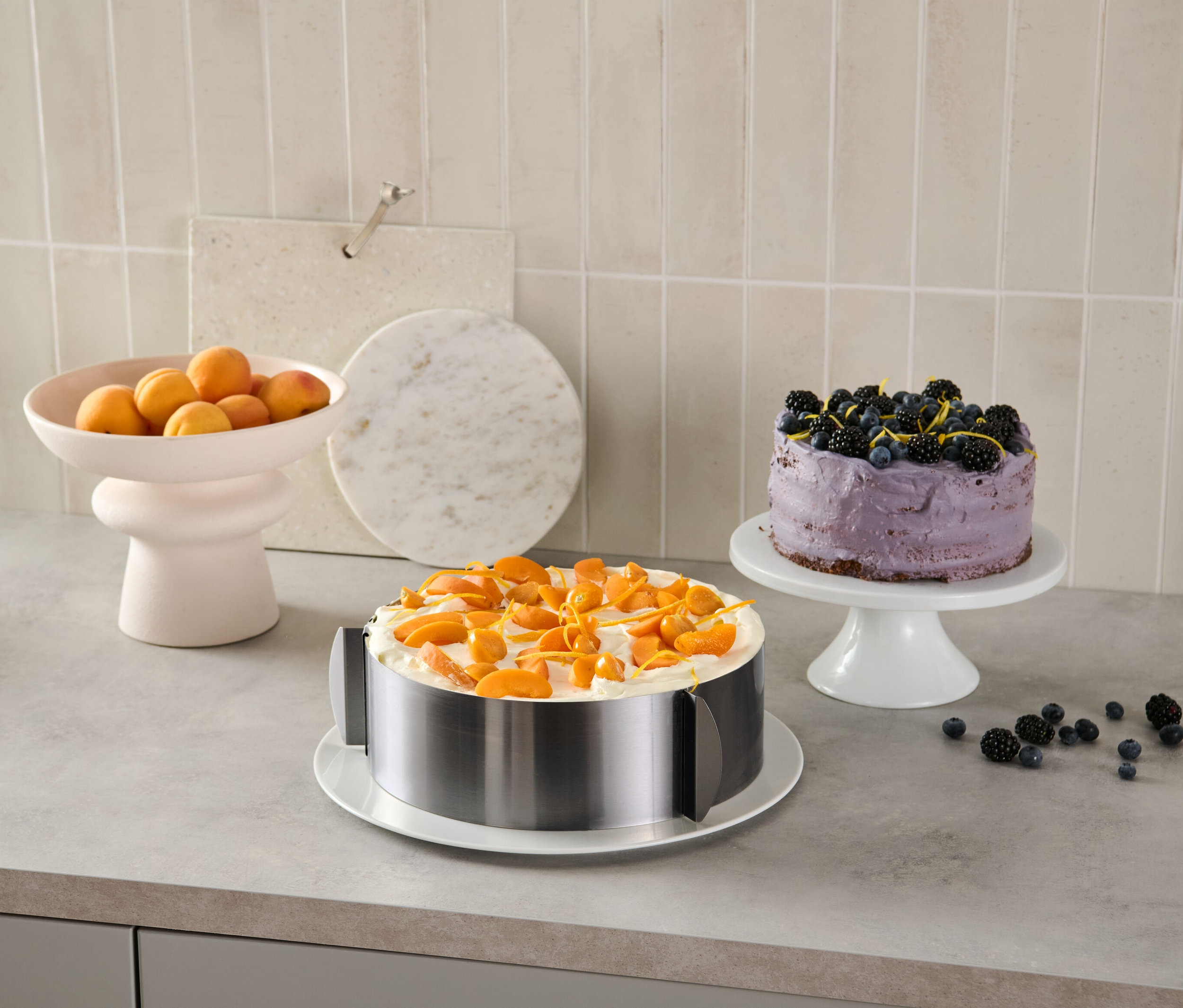 Auf einer Küchentheke steht ein mit Aprikosen dekorierter Kuchen in einem Edelstahl-Tortenring, im Hintergrund ein Blaubeerkuchen und eine Schale mit Aprikosen.