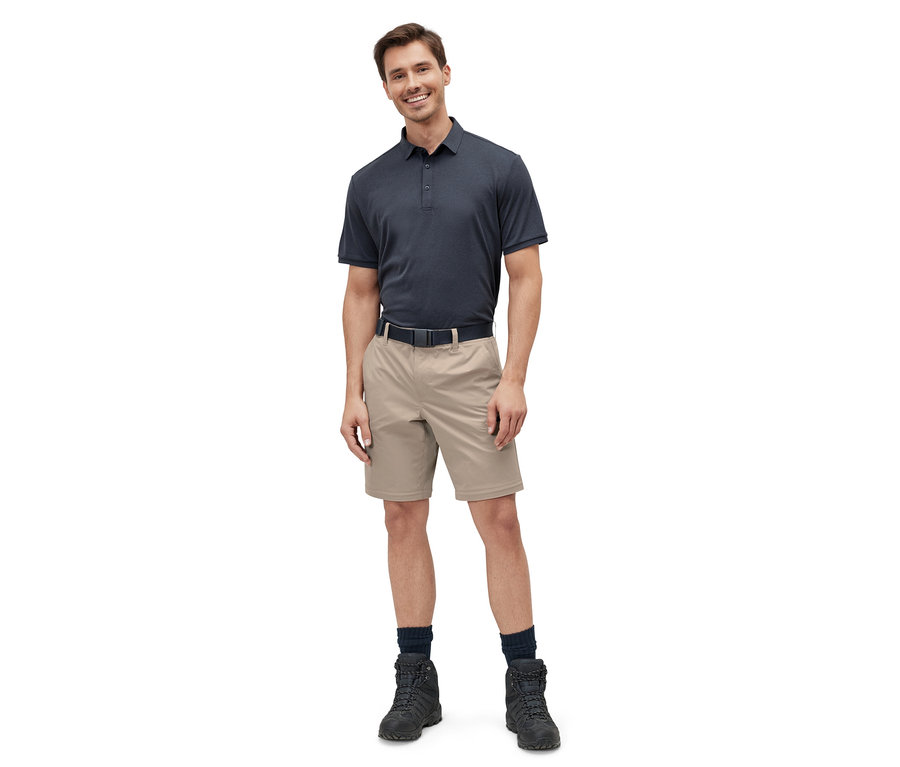 Mann steht und trägt eine beige 2-in-1-Funktionshose, ein marineblaues Poloshirt, einen schwarzen Gürtel und schwarze Stiefel.